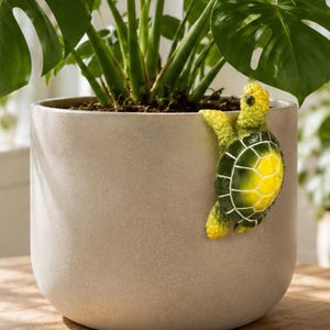 May include: A light grey ceramic plant pot with a decorative turtle climbing on the side. The turtle has a yellow and green shell. The pot contains a green plant with large leaves.