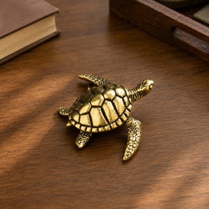 May include: A detailed, golden-colored turtle figurine. The turtle has a textured shell and detailed legs and head. The figurine is placed on a wooden surface, suggesting a decorative or collectible item.