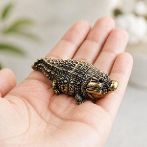 May include: A detailed, antique-style brass alligator figurine. The sculpture features textured scales and a detailed head, showcasing a vintage aesthetic. The alligator is a decorative item, likely a collectible or a piece of home decor.