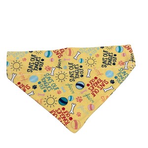 Peut inclure: Un bandana triangulaire jaune pour animal de compagnie avec un motif répétitif de soleils, de balles colorées, d'os blancs, d'empreintes de pattes, du texte "SUN'S OUT TONGUES OUT" en bleu foncé et rouge, et le mot "woof" en écriture cursive verte.
