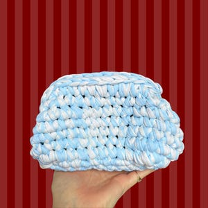 May include: A handmade, crocheted clutch in a mix of light blue and white. The bag is held in a hand, showcasing the texture and color pattern. The background is a red and white striped pattern.