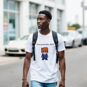 Könnte beinhalten: Weißes T-Shirt mit dem Text "Corporate NPC" und einer pixeligen Figur mit braunen Haaren und einem blauen Hemd. Die Person trägt einen schwarzen Rucksack und geht auf einer Straße.