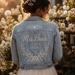 Könnte beinhalten: Hellblaue Jeansjacke mit weißer Blumenstickerei und dem Text "Mrs. Bruce" über einem Herz mit dem Datum "6.7.20". Die Jacke ist Teil eines Brautensembles.