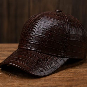 Peut inclure: Casquette de baseball en cuir marron foncé avec un motif texturé en forme de crocodile. La casquette a une visière incurvée et un bouton sur le dessus. Le chapeau est conçu pour être porté à l'extérieur.