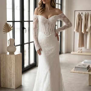 Peut inclure: Une robe de mari&eacute;e longue en dentelle blanche avec une encolure d&eacute;gag&eacute;e, des manches longues en dentelle transparente et un corsage structur&eacute; style corset orn&eacute; de perles d&eacute;licates le long de l'encolure et des coutures du bustier.
