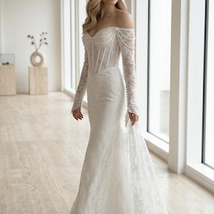Peut inclure: Une robe longue en dentelle blanche avec une encolure d&eacute;gag&eacute;e, des manches longues et un corsage structur&eacute; style corset. Le v&ecirc;tement est orn&eacute; de d&eacute;licats rangs de perles drap&eacute;s sur la poitrine et les &eacute;paules, avec une tra&icirc;ne.