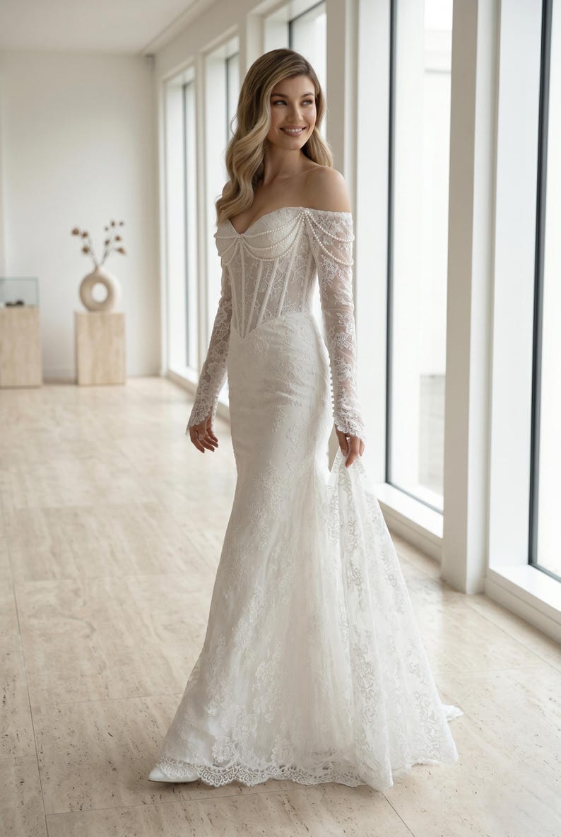 Peut inclure: Une robe longue en dentelle blanche avec une encolure d&eacute;gag&eacute;e, des manches longues et un corsage structur&eacute; style corset. Le v&ecirc;tement est orn&eacute; de d&eacute;licats rangs de perles drap&eacute;s sur la poitrine et les &eacute;paules, avec une tra&icirc;ne.