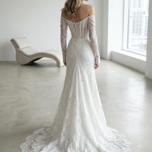 Peut inclure: Une robe de mari&eacute;e blanche en dentelle longue jusqu'au sol avec une encolure d&eacute;gag&eacute;e, des manches longues et un corsage style corset avec un la&ccedil;age au dos. La robe pr&eacute;sente une coupe ajust&eacute;e qui s'&eacute;vase en une tra&icirc;ne &agrave; bordure festonn&eacute;e.