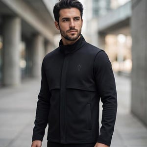 Puede incluir: Chaqueta negra con cuello alto y un pequeño emblema en el pecho. La chaqueta tiene un diseño aerodinámico con cremallera frontal y bolsillos. La persona está de pie afuera frente a un edificio.