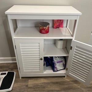 May include: A white wooden cabinet with a slatted door and open shelves. The cabinet contains a candle, toilet paper, and a bag of bath salts. A digital scale is on the floor next to the cabinet.