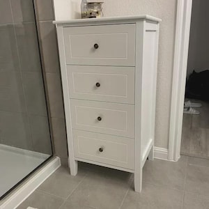 May include: A white, four-drawer cabinet with dark knobs. The cabinet is rectangular and stands on four legs. A glass jar and a bottle sit on top. The cabinet is in a bathroom setting.
