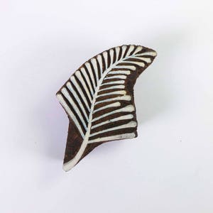 May include: A dark brown wooden printing block featuring a leaf design. The leaf pattern is white with light blue accents, resembling a fern. This block is likely used for textile printing or crafting, with a curved shape.