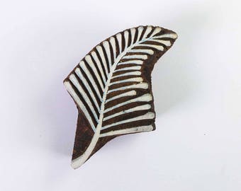 May include: A dark brown wooden printing block featuring a leaf design. The leaf pattern is white with light blue accents, resembling a fern. This block is likely used for textile printing or crafting, with a curved shape.