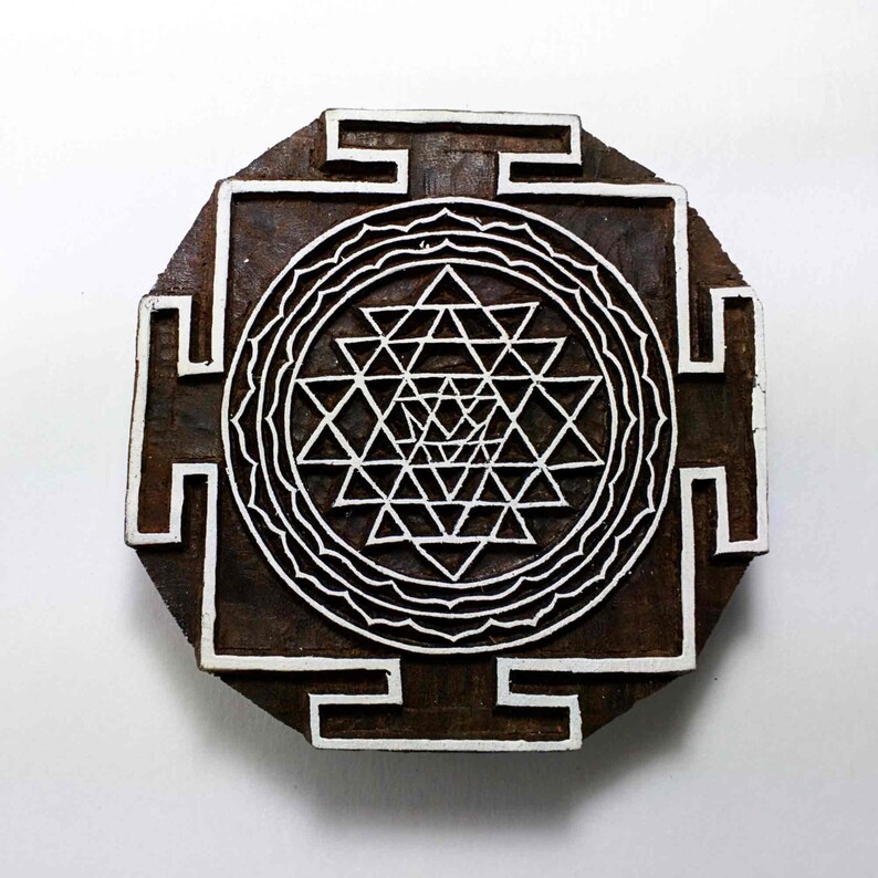 May include: A wooden block printing stamp with a white Sri Yantra design. The design is a geometric pattern of interlocking triangles within a circle.