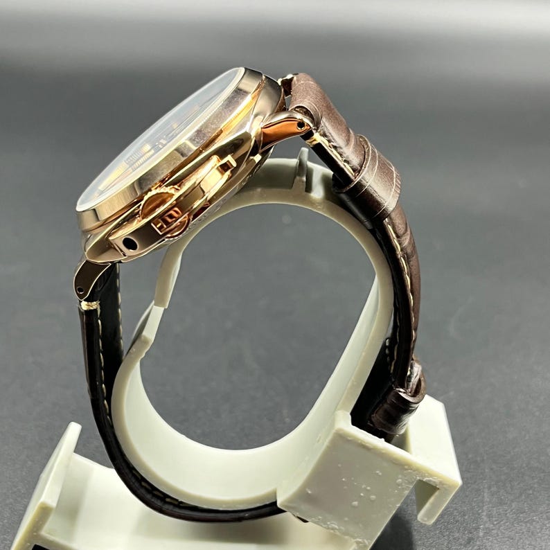 Puede incluir: Un reloj de pulsera con caja de metal en tono oro rosa pulido y correa de cuero marr&oacute;n oscuro con acabado texturizado y costuras blancas. El lateral de la caja muestra un protector de corona prominente y una corona texturizada para el ajuste.