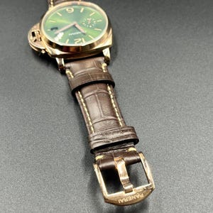 Puede incluir: Un reloj de pulsera Panerai con caja redonda en tono dorado, esfera verde con efecto rayos de sol, manecillas e &iacute;ndices dorados, y una correa de cuero marr&oacute;n con textura de cocodrilo, costuras en contraste y hebilla dorada.
