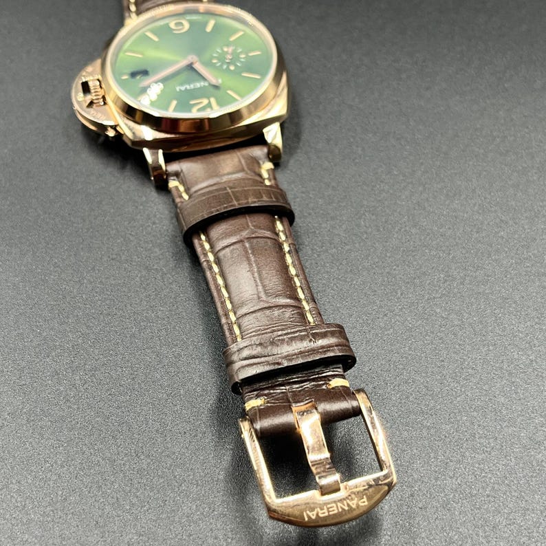 Puede incluir: Un reloj de pulsera Panerai con caja redonda en tono dorado, esfera verde con efecto rayos de sol, manecillas e &iacute;ndices dorados, y una correa de cuero marr&oacute;n con textura de cocodrilo, costuras en contraste y hebilla dorada.