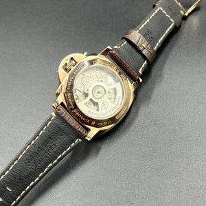 Puede incluir: Un reloj Officine Panerai Luminor Composite de tono dorado con una parte posterior transparente que revela el movimiento mec&aacute;nico interno. El reloj cuenta con una correa de cuero marr&oacute;n oscuro con costuras blancas y est&aacute; marcado Made in Austria.