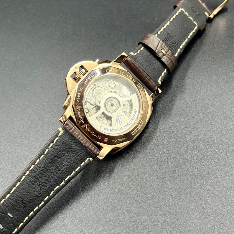 Puede incluir: Un reloj Officine Panerai Luminor Composite de tono dorado con una parte posterior transparente que revela el movimiento mec&aacute;nico interno. El reloj cuenta con una correa de cuero marr&oacute;n oscuro con costuras blancas y est&aacute; marcado Made in Austria.