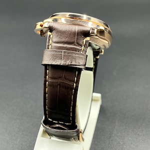 Puede incluir: Un reloj de pulsera con caja de metal en tono oro rosa pulido y correa de cuero marr&oacute;n oscuro con un patr&oacute;n texturizado de imitaci&oacute;n de cocodrilo. La correa est&aacute; asegurada con costuras claras visibles en los bordes y una hebilla de metal a juego.