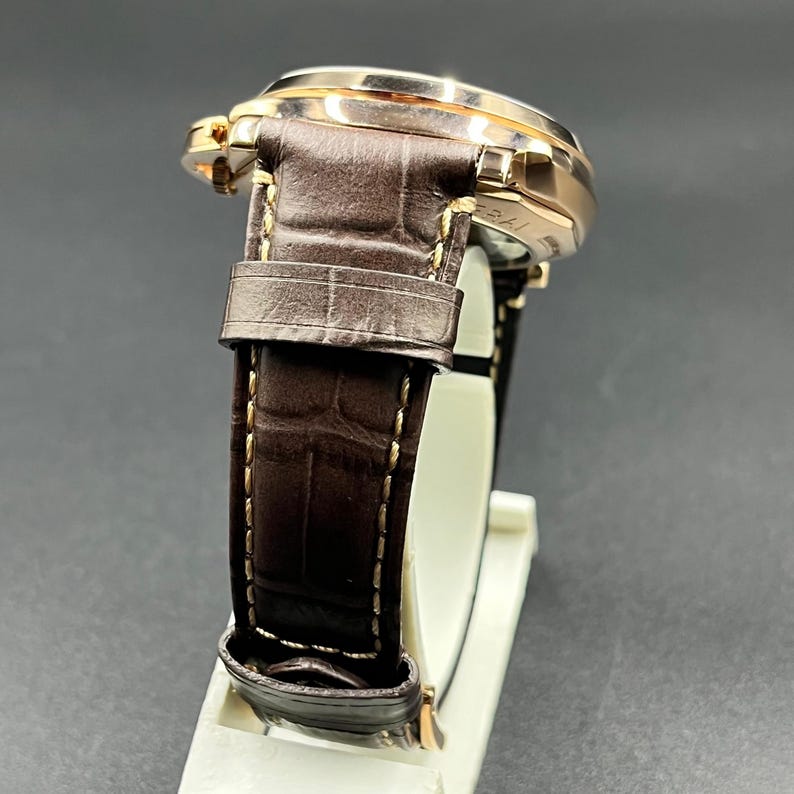 Puede incluir: Un reloj de pulsera con caja de metal en tono oro rosa pulido y correa de cuero marr&oacute;n oscuro con un patr&oacute;n texturizado de imitaci&oacute;n de cocodrilo. La correa est&aacute; asegurada con costuras claras visibles en los bordes y una hebilla de metal a juego.