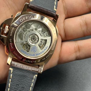Puede incluir: Un reloj Officine Panerai Luminor Composite en tono oro rosa con fondo de caja transparente que muestra el movimiento mec&aacute;nico. La parte posterior tiene grabados como Officine Panerai, Luminor Composite y 300m, con una correa de cuero oscuro.