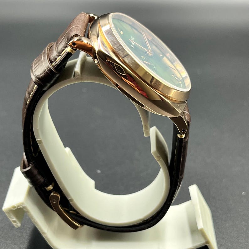 Puede incluir: Un reloj de pulsera con caja de tono oro rosa pulido y esfera verde oscuro con manecillas e &iacute;ndices dorados. El reloj cuenta con una correa de cuero texturizado en color marr&oacute;n oscuro que incluye una hebilla met&aacute;lica a juego en oro rosa.