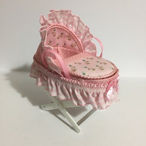 May include: A miniature pink baby cradle with a frilly, ruffled trim. The cradle features a patterned interior with small animal figures and is set on a white, folding stand. A decorative bow adorns the side.