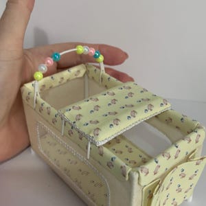 May include: A miniature dollhouse bassinet with a yellow patterned fabric featuring small animal figures. The bassinet has a cream-colored exterior, a beaded handle, and a hinged canopy. The overall design is soft and pastel-colored.