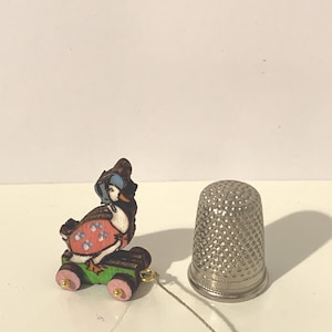 Könnte beinhalten: Eine Miniatur-Holzente-Figur mit Hut und Kleid, die auf einem grünen und braunen Wagen mit rosa Rädern sitzt. Ein silberner Fingerhut befindet sich neben der Ente. Die Entenfigur ist mit einem dünnen, silbernen Faden verbunden.