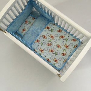 May include: A white wooden crib with blue bedding. The bedding includes a bumper, a small pillow, and a blanket. The blanket and pillow feature a light blue pattern with Winnie the Pooh characters, clouds, and umbrellas. The crib is empty.