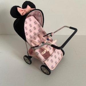 Può includere: Un passeggino rosa e nero in miniatura a tema Minnie Mouse. Il passeggino ha un cappuccio nero con orecchie di Minnie Mouse e un fiocco rosa. Il sedile è rosa con un motivo ripetuto di Minnie Mouse. Il passeggino ha quattro piccole ruote e un manico argentato.