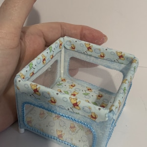 May include: A blue and white Winnie the Pooh-themed playpen with a mesh side. The playpen has a Winnie the Pooh print with clouds and umbrellas.