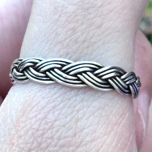 May include: A silver-toned ring featuring a continuous braided pattern that wraps around the entire band. The metal has a polished finish with darkened recessed areas that highlight the intricate woven texture of the design.