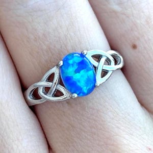 May include: A silver-toned ring featuring a central oval-shaped blue opal stone with iridescent highlights. The band is crafted with an intricate, open-work knot design on both sides of the stone, creating a continuous pattern around the finger.