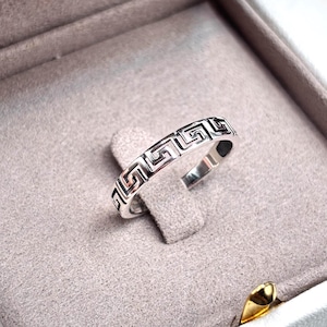 May include: A silver ring with a Greek key pattern is displayed inside a white jewelry box with a gray interior. The ring is centered and the box is open, showcasing the ring's design.
