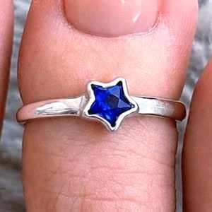 May include: A silver-toned toe ring featuring a small, five-pointed star-shaped setting. The center of the star contains a faceted, deep blue gemstone. The band of the ring is smooth, polished, and metallic, encircling the toe with a simple design.
