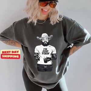 May include: A charcoal gray short-sleeve t-shirt featuring a black and white graphic of a bearded person wearing a cowboy hat and a white t-shirt that says I'M JUST VIBIN' while holding a drink. The graphic also shows tattoos on the arms.