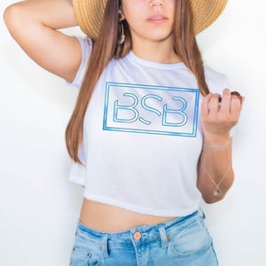 May include: A white short-sleeved crop top featuring a rectangular graphic on the chest. The graphic contains the letters BSB in a light blue outline style, enclosed within a matching light blue rectangular border against a white background.