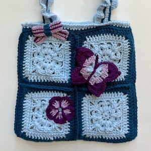 May include: A handmade crochet tote bag featuring a patchwork of light blue and dark blue granny squares. The bag is adorned with a purple and light blue butterfly appliqué, a purple flower, and a small striped bow. It has a ruffled light blue handle.
