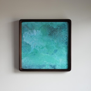 May include: A square wall art piece featuring a textured, abstract surface in shades of turquoise, teal, and deep blue. The artwork is encased in a dark brown wooden frame with rounded corners, creating a clean, defined border around the painting.