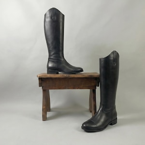 以下が含まれることがあります： 黒い革製の乗馬ブーツが2足。1足は小さな木製のスツールの上に、もう1足は床に置かれています。ブーツは背が高く、クラシックなデザインで、滑らかな仕上がりです。ブーツにはわずかなヒールがあります。