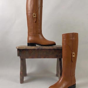 Puede incluir: Un par de botas de montar de cuero color canela hasta la rodilla con acabado liso, tacón bajo y un detalle de hebilla de metal dorado en el lado exterior de cada caña. Las botas tienen una silueta estructurada con costuras verticales.