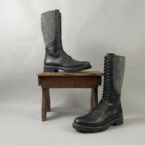 May include: A pair of black leather knee-high boots with grey suede panels. The boots feature a lace-up design and are displayed on a small wooden stool. The soles are thick and rugged.