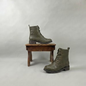 May include: Olive green lace-up boots with a low heel. One boot is on a small, rustic wooden stool, and the other is on the floor. The boots have a classic combat boot style.