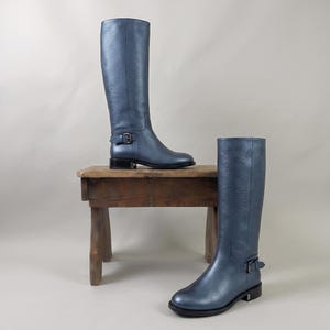 Op de afbeelding: Een paar kniehoge laarzen in een metallic blauwe afwerking met een getextureerd oppervlak. Elke laars heeft een ronde neus, een lage blokhak en een decoratieve band over de enkel, vastgezet met een donkere metalen gesp en bijpassend beslag.