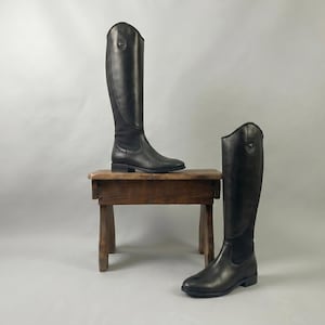 May include: Two black leather riding boots. One boot is on a small, rustic wooden stool, while the other stands on the floor. The boots have a tall, sleek design, with a slight heel and a smooth finish. The background is a plain, light gray.