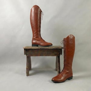 May include: A pair of tall, brown leather riding boots with lace-up fronts. One boot is on a small, weathered wooden stool, while the other rests on the floor. The boots have a classic equestrian design.