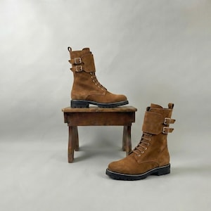 May include: Two brown suede combat boots with black soles and buckle details. One boot is on a small wooden stool, the other is on the floor. The boots have laces and a mid-calf height.