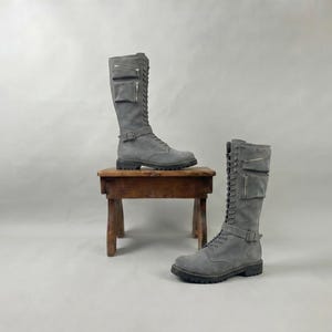 May include: Two grey suede knee-high boots with lace-up fronts, side zips, and buckle details. One boot rests on a small wooden stool, the other on the floor. The boots have a rugged, yet stylish design, suitable for various outfits.